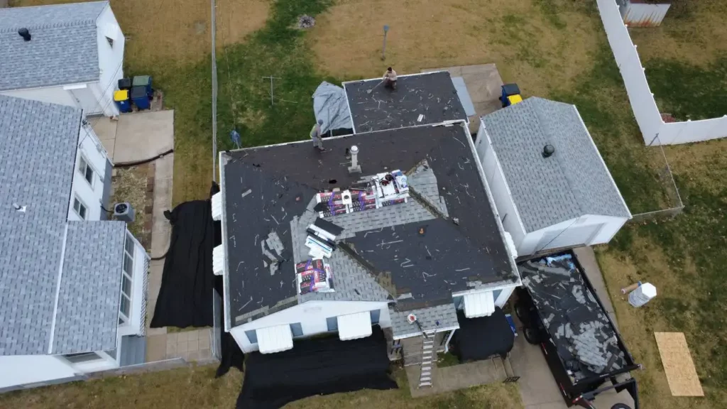 An aerial view of a house undergoing roof repairs, with workers on the roof and construction materials scattered around. Black tarps cover parts of the yard, and surrounding houses are visible.