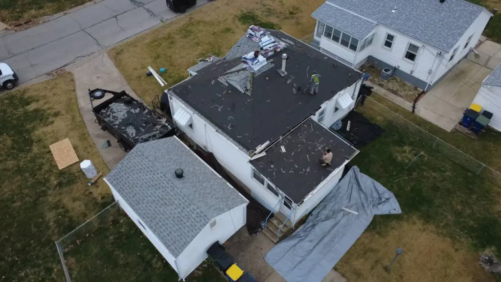An aerial view of a house with roofing work in progress. Workers are seen on the roof, surrounded by materials and debris. Tarps are laid out on the ground, with some roofing shingles visible.