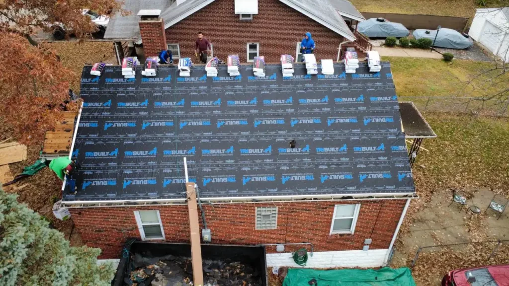 Aerial view of a house with a newly installed roof covered in black underlayment. Several workers can be seen on the roof preparing for shingles. The yard is filled with fallen leaves, and there are tarps and tools scattered below.