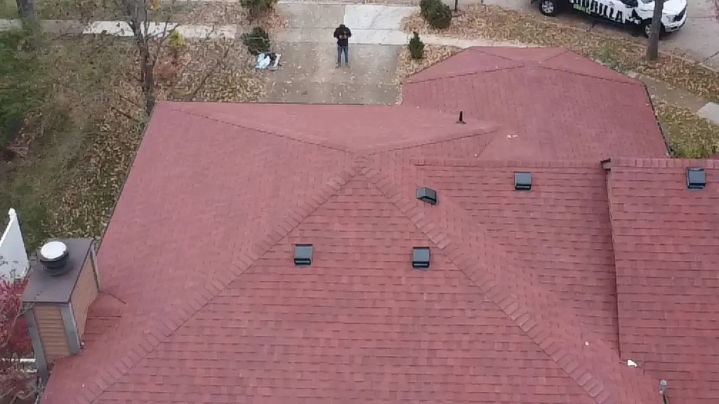 Aerial view of a red sloped roof with ventilation openings. In the background, a person stands on a sidewalk surrounded by autumn leaves, with vehicles parked nearby.