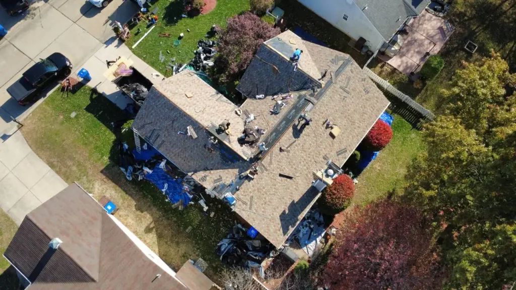 An aerial view of a house under renovation, showing construction workers on the roof and various materials scattered in the yard. The landscape features green grass, flowering trees, and visible vehicles parked nearby.